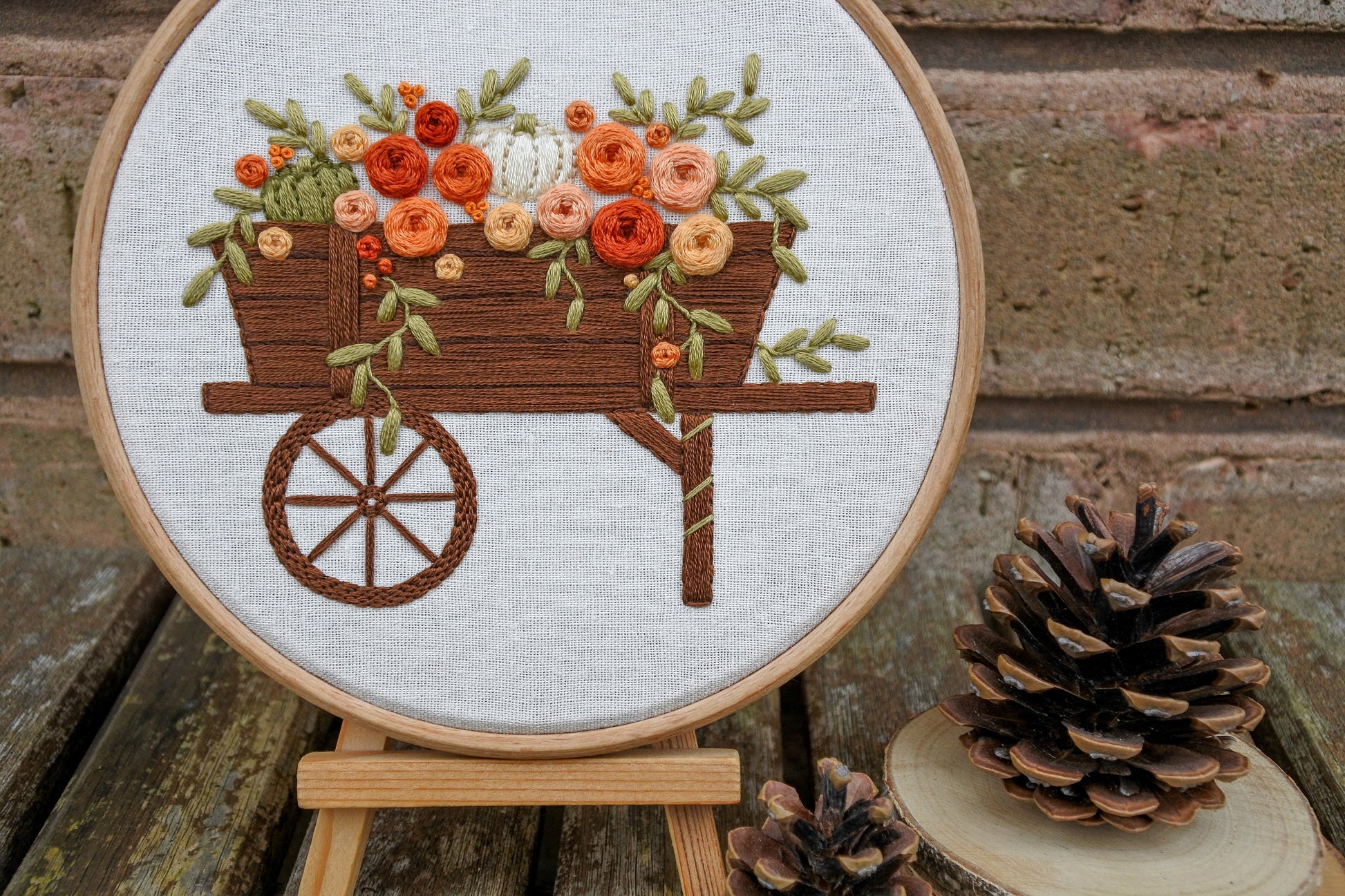 Autumn floral pumpkin wagon embroidery displayed in a wooden hoop on a mini wooden easel next to pinecones