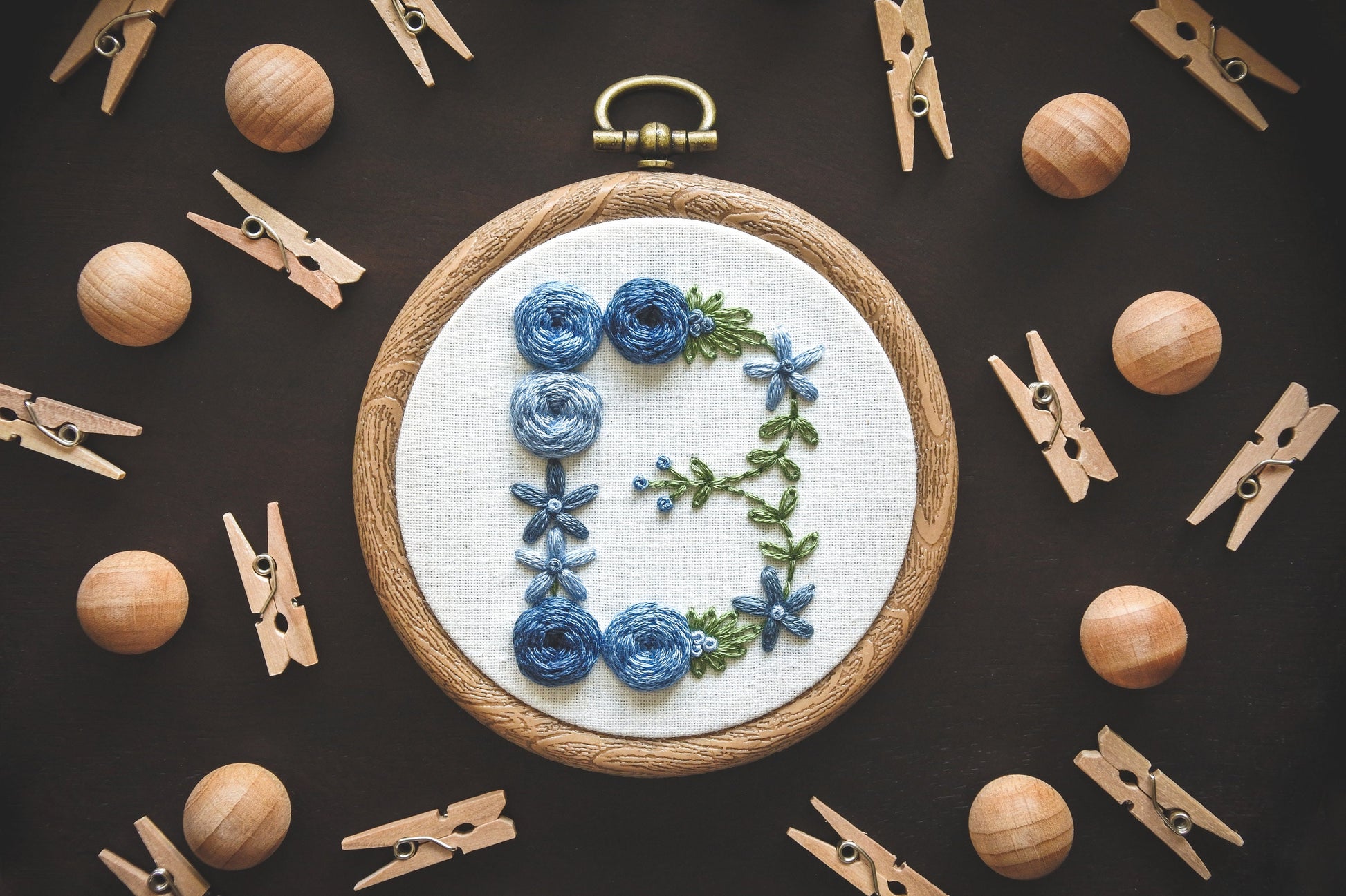 Hand-embroidered blue floral letter 'B' hoop with green foliage. Decorative background of mini wooden pegs and balls.