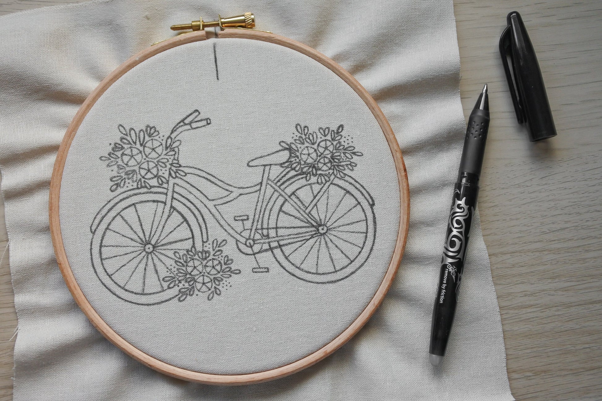 Floral bicycle embroidery pattern transferred onto neutral cotton fabric with a heat erasable pen placed beside the hoop.