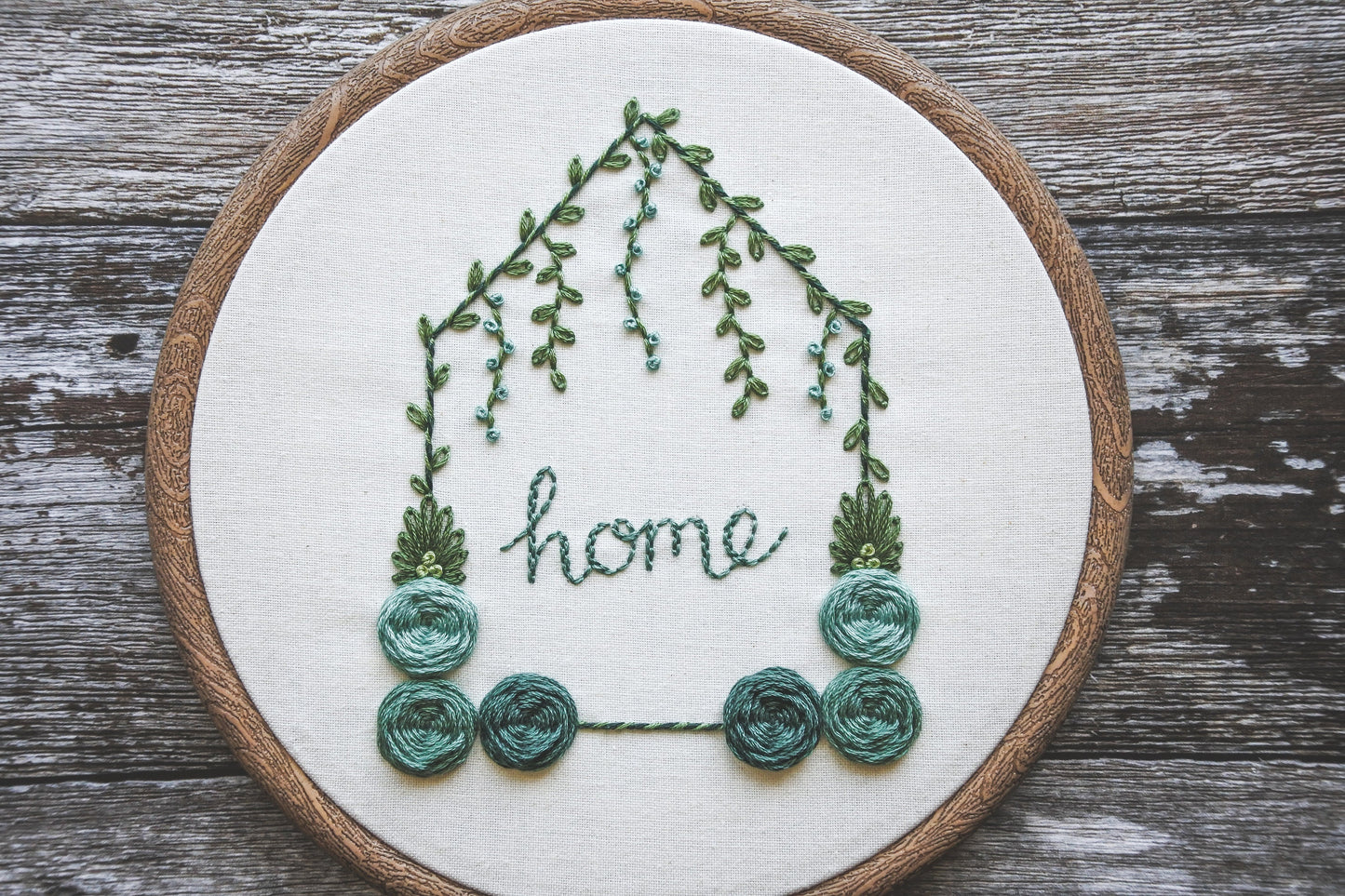 Hand-embroidered floral home hoop with turquoise colour scheme, green foliage, and the word 'home' in the centre.