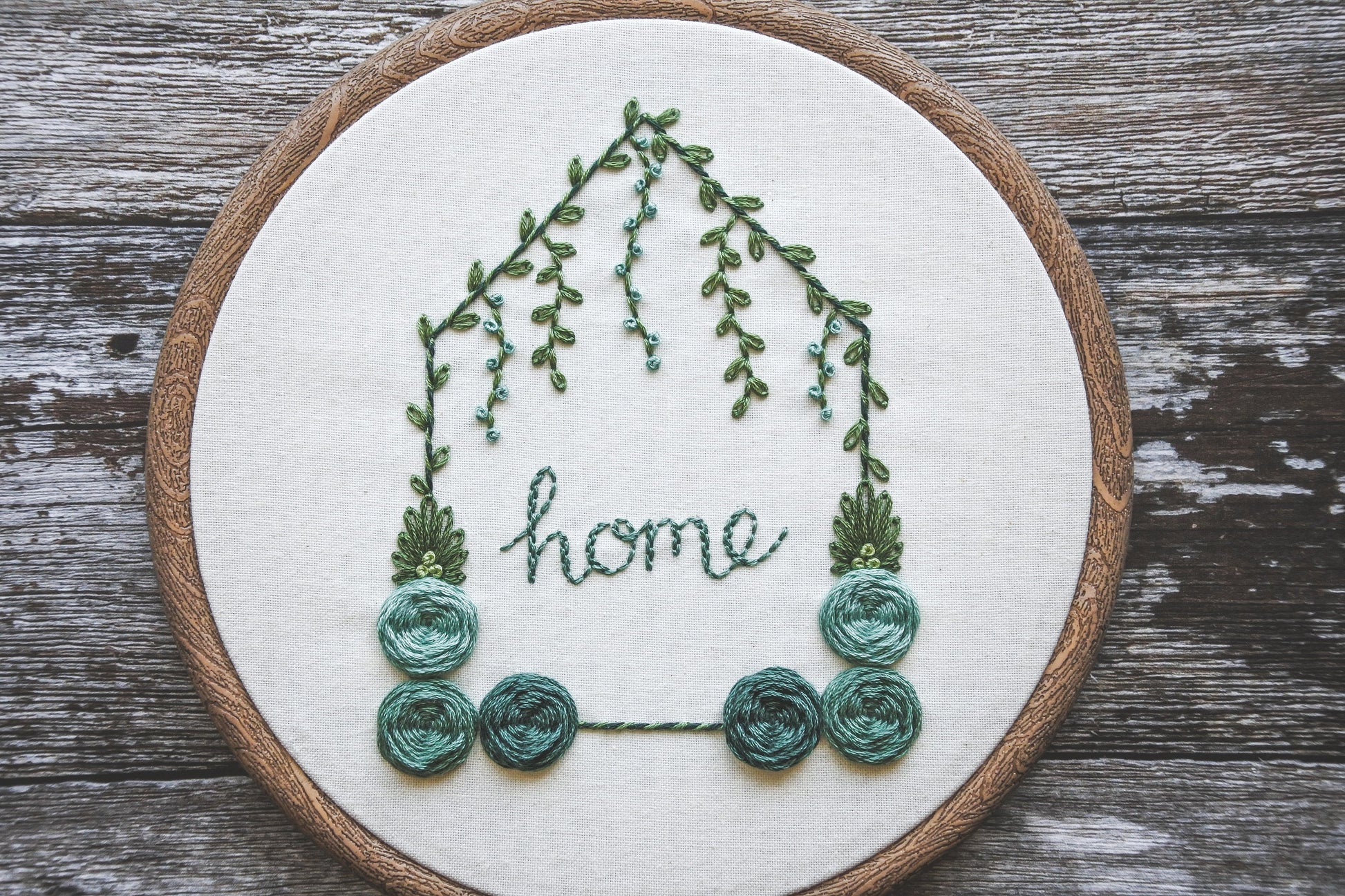 Hand-embroidered floral home hoop with turquoise colour scheme, green foliage, and the word 'home' in the centre.