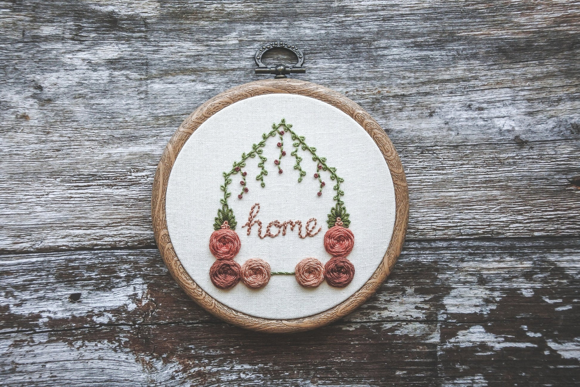 Hand-embroidered floral home hoop with red and peach colours, green foliage, and the word 'home' in the centre, in a wood effect hoop frame.