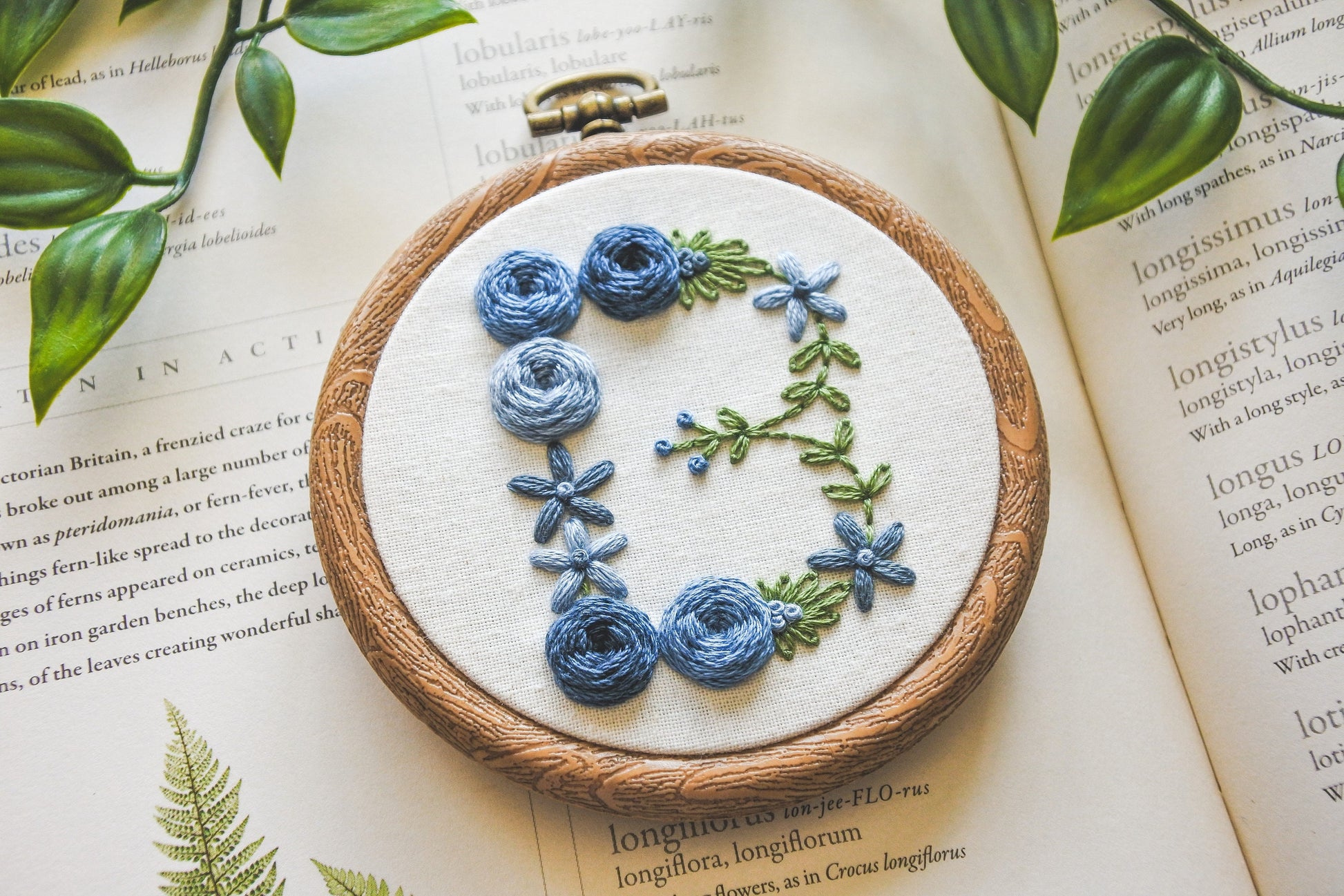 Closeup of floral letter B hand embroidery hoop with muted blue flowers and green foliage.