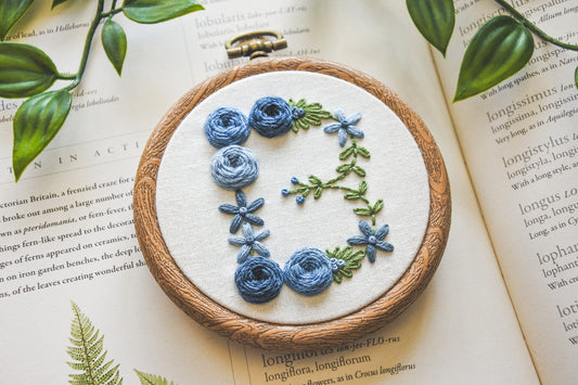 Closeup of floral letter B hand embroidery hoop with muted blue flowers and green foliage.