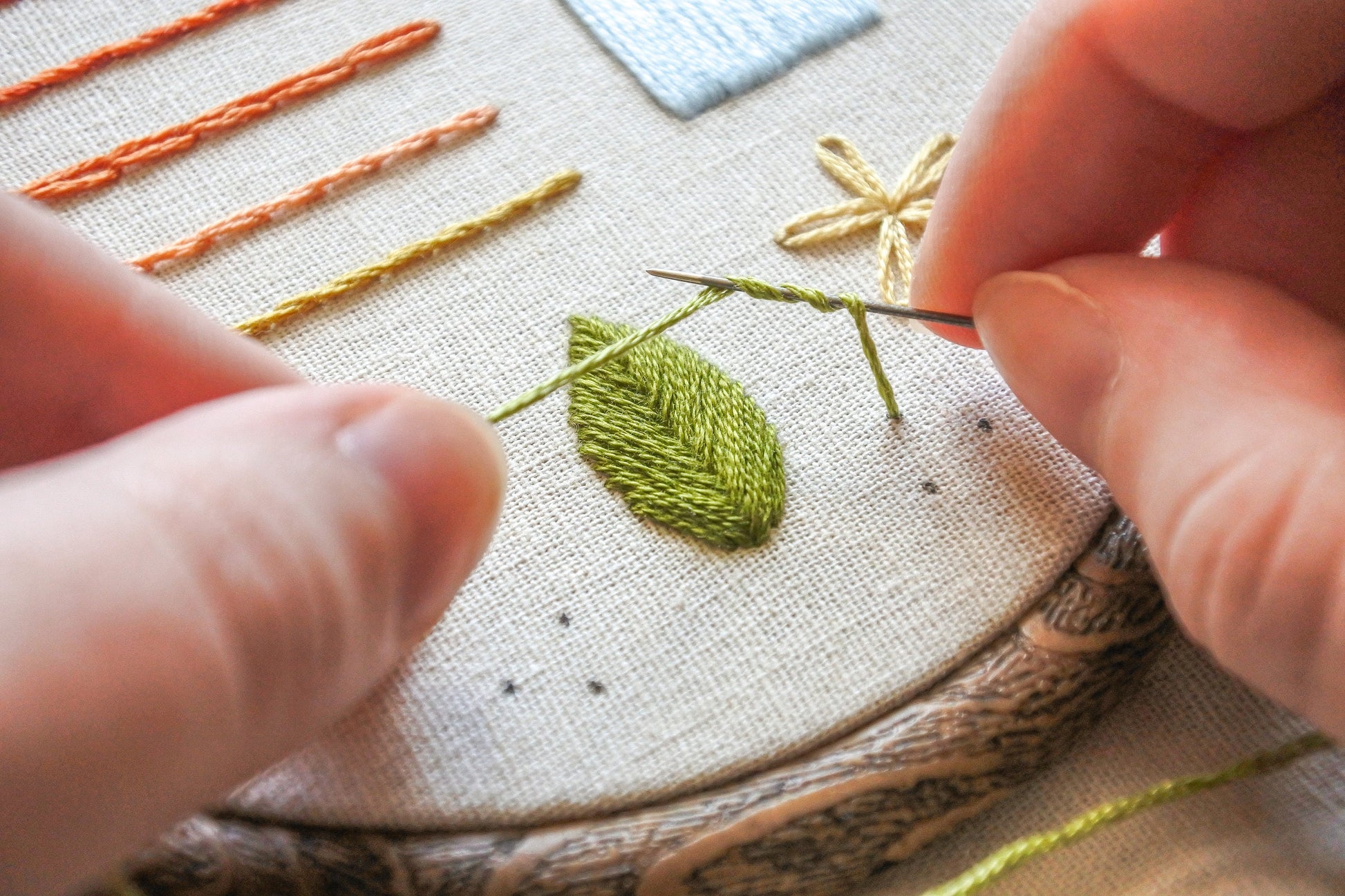 Hands holding a needle and thread in the process of stitching a french knot with green thread on light neutral fabric.