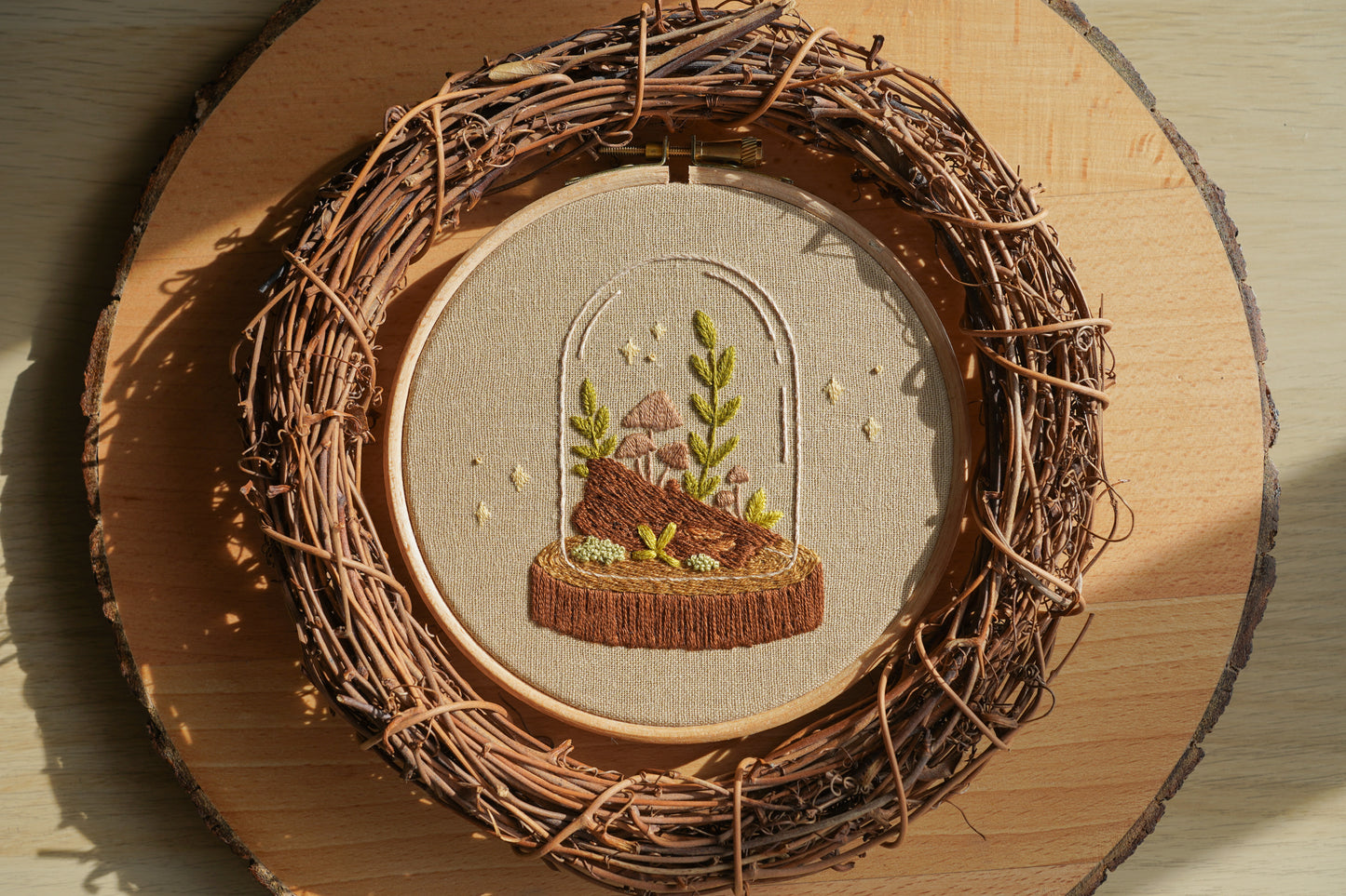 Sunlight shining on embroidery of a terrarium with plants and a mushroom inside a wooden hoop, surrounded by a twig wreath on a log board.