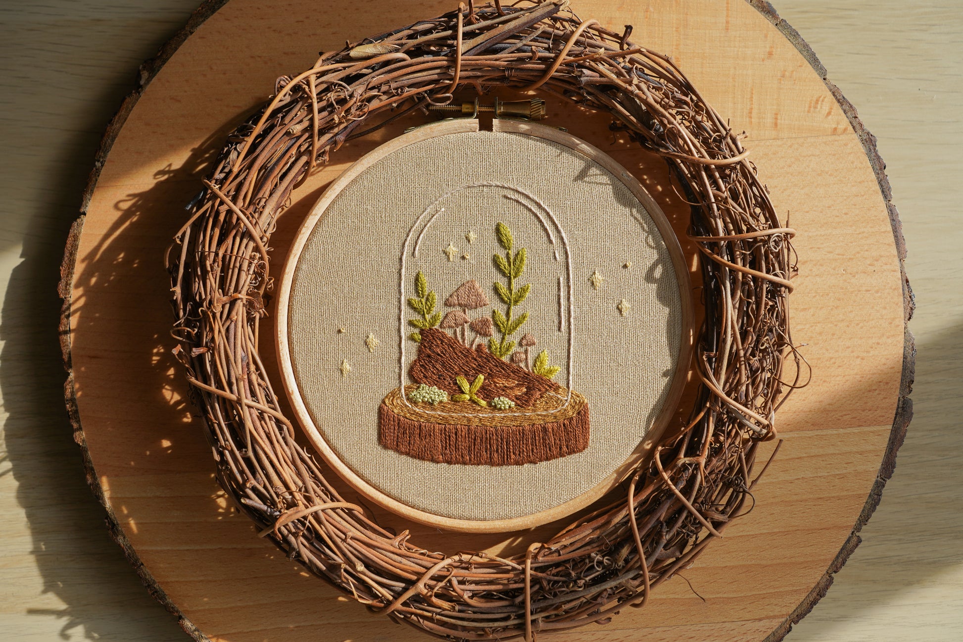 Sunlight shining on embroidery of a terrarium with plants and a mushroom inside a wooden hoop, surrounded by a twig wreath on a log board.
