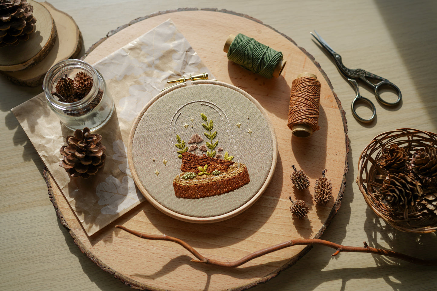 Embroidery hoop with nature-themed woodland mushroom terrarium design on a wooden surface with embroidery supplies.