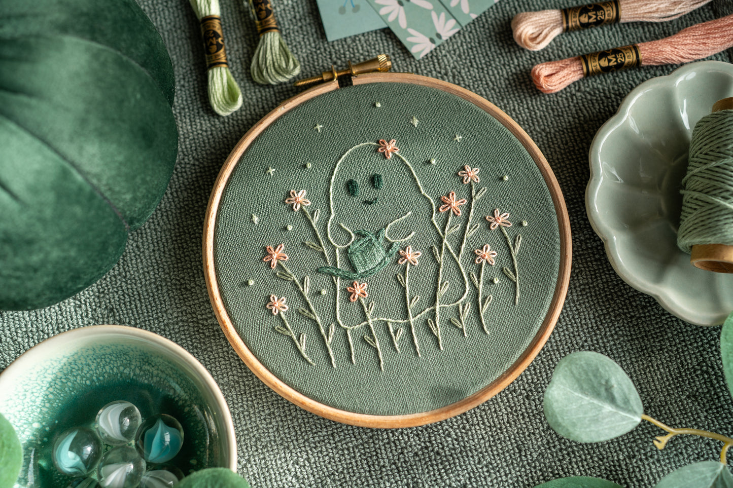 Hand embroidered ghost watering pink flowers with a blue watering can on sage green fabric in a wooden embroidery hoop. 