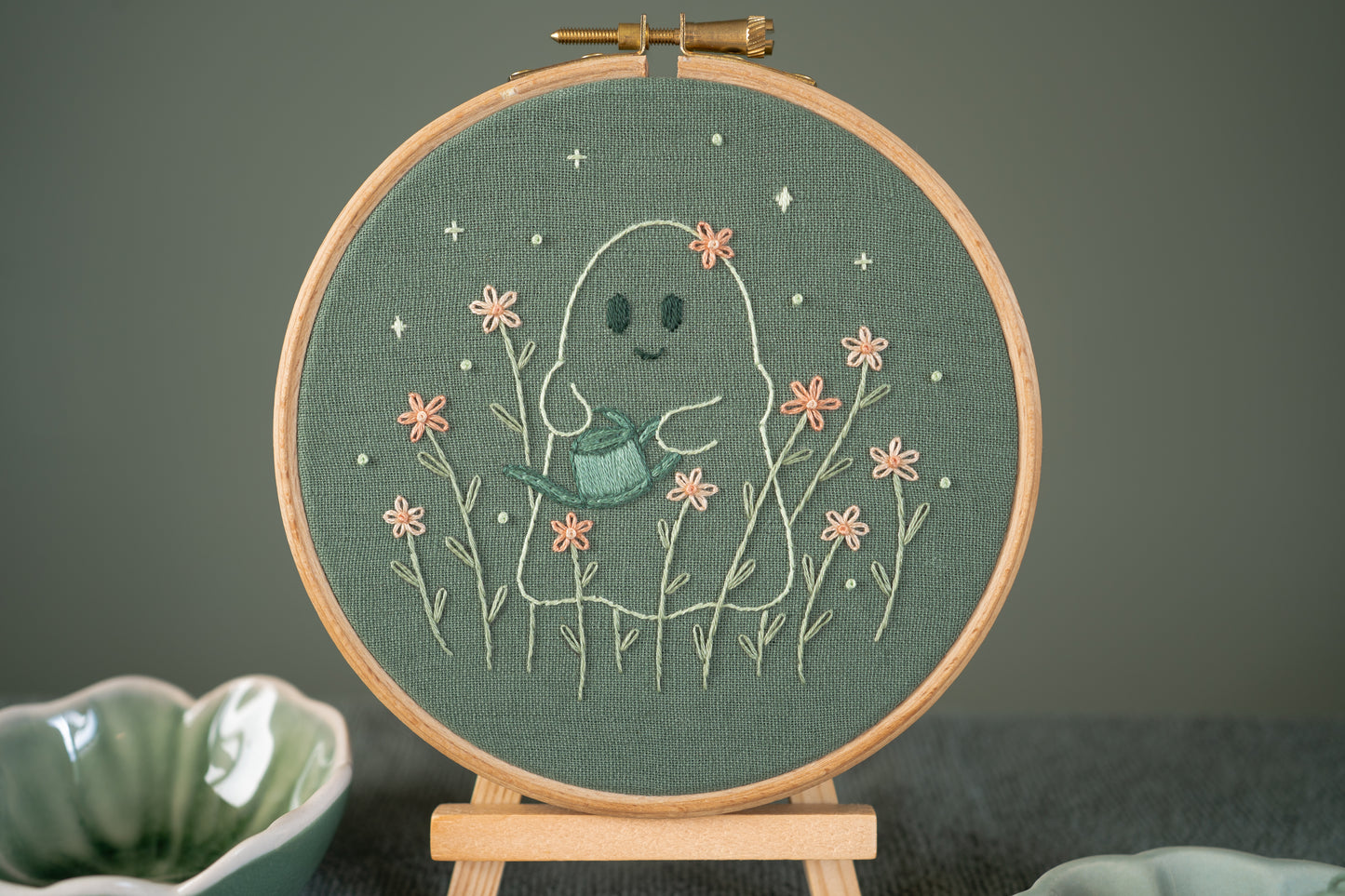 Ghost gardener hand embroidery design on sage green fabric in a wooden hoop displayed on a mini wooden easel.