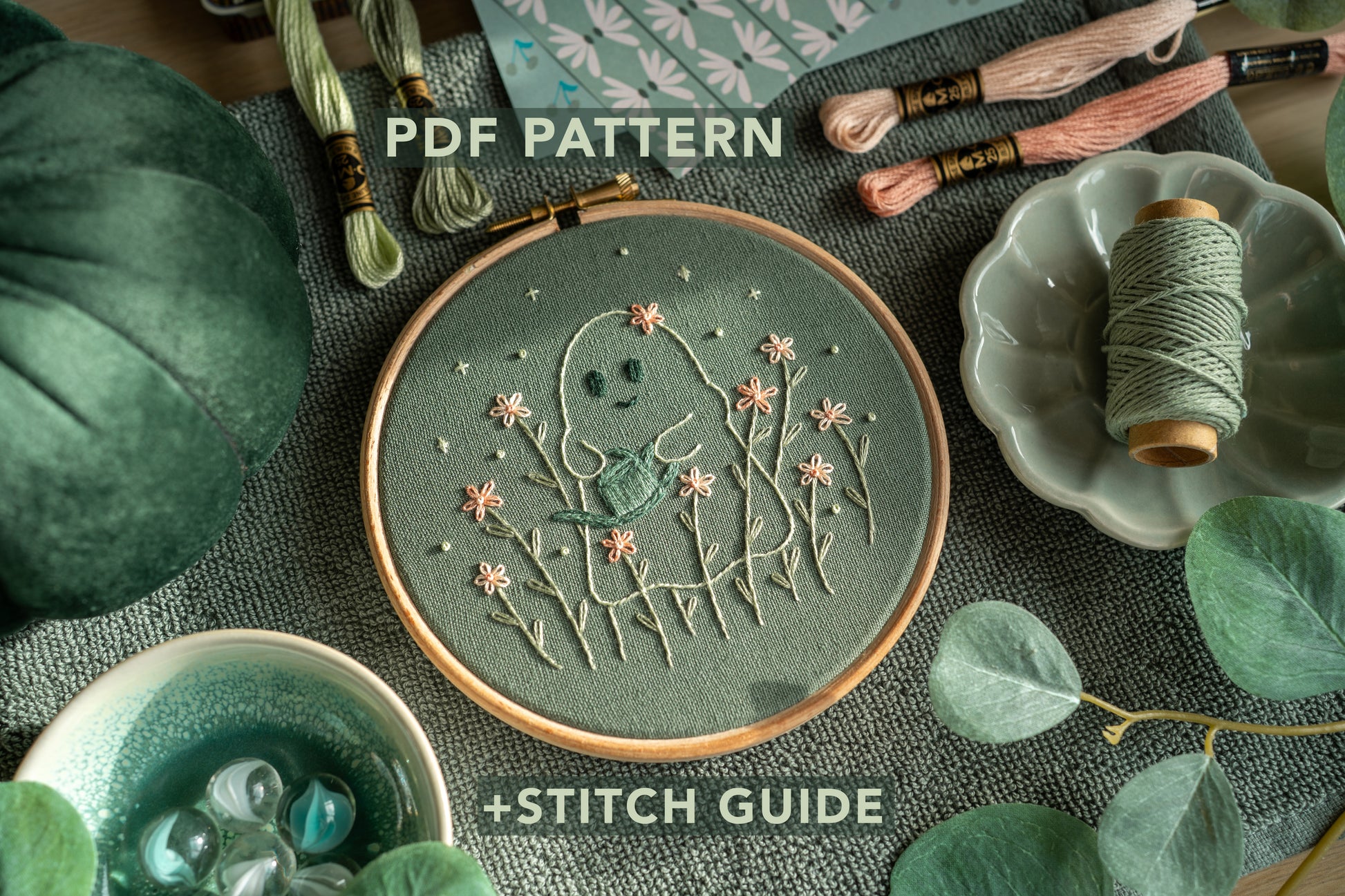 Hand embroidery of a happy ghost holding a blue watering can, surrounded by pink flowers, stitched on sage green fabric in a wooden embroidery hoop, with text overlay reading “PDF pattern and stitch guide”