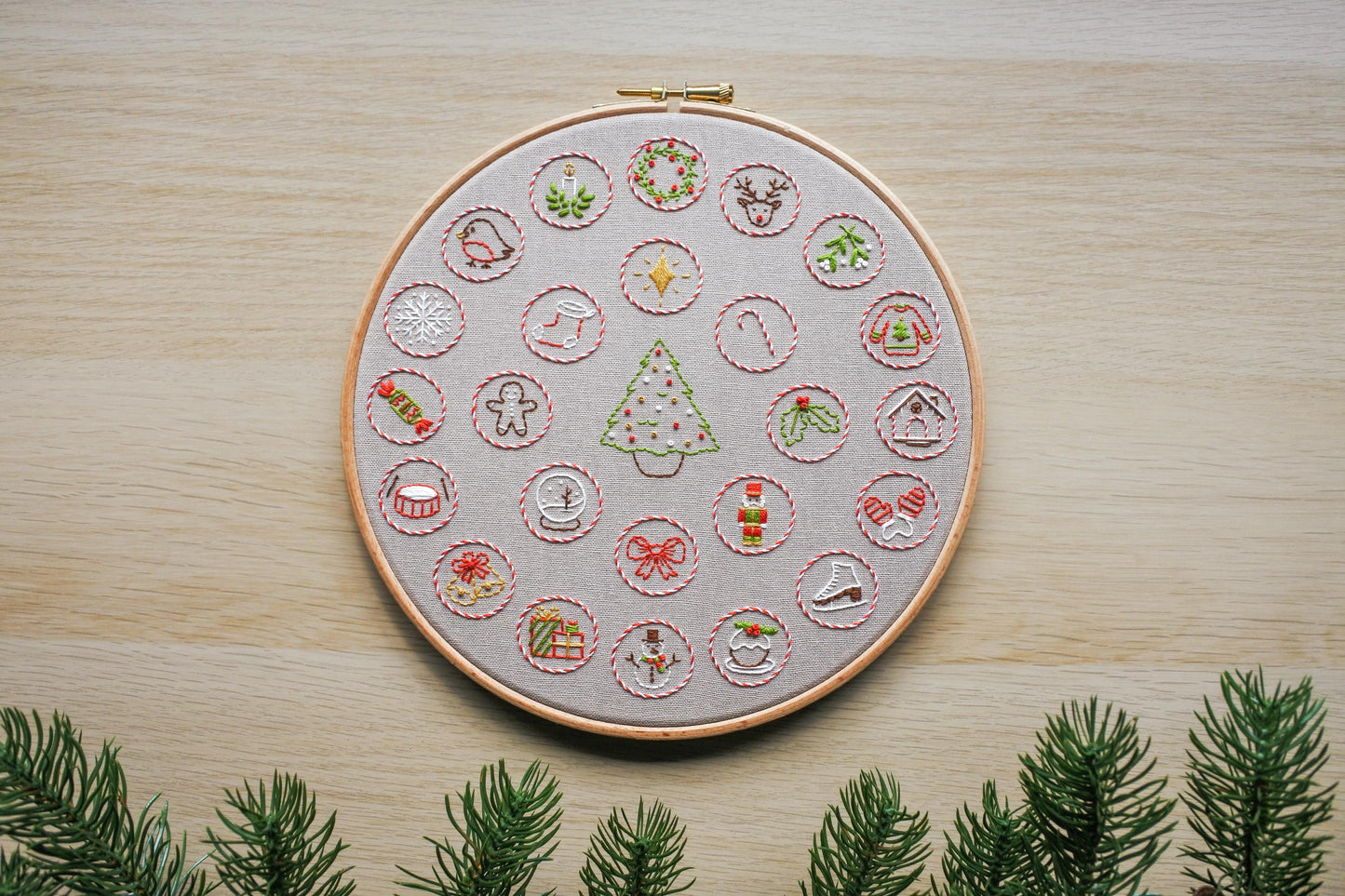 Hand embroidered advent calendar, stitched on light beige fabric, featuring 24 mini Christmas themed designs in striped candy cane red and white circles surrounding a Christmas tree.