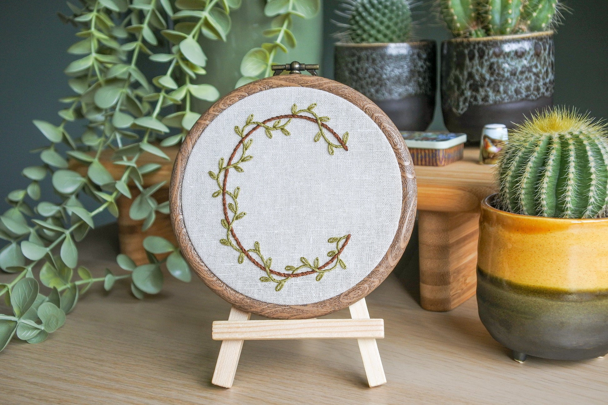 Hand embroidered letter C with green vines and brown thread on light neutral fabric in a wood-effect hoop, displayed on a mini wooden easel.