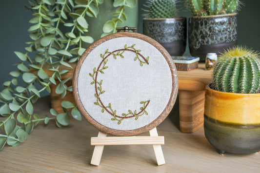 Hand embroidered letter C with green vines and brown thread on light neutral fabric in a wood-effect hoop, displayed on a mini wooden easel.