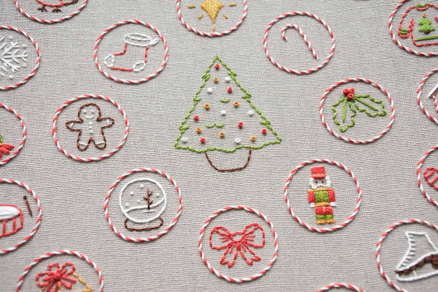 Closeup of hand embroidered christmas tree on light beige fabric; surrounded by red and white candy can striped circles, each featuring a mini festive stitched design including a snow globe, holly leaves, gingerbread man, nutcracker doll, bow, stocking and more!