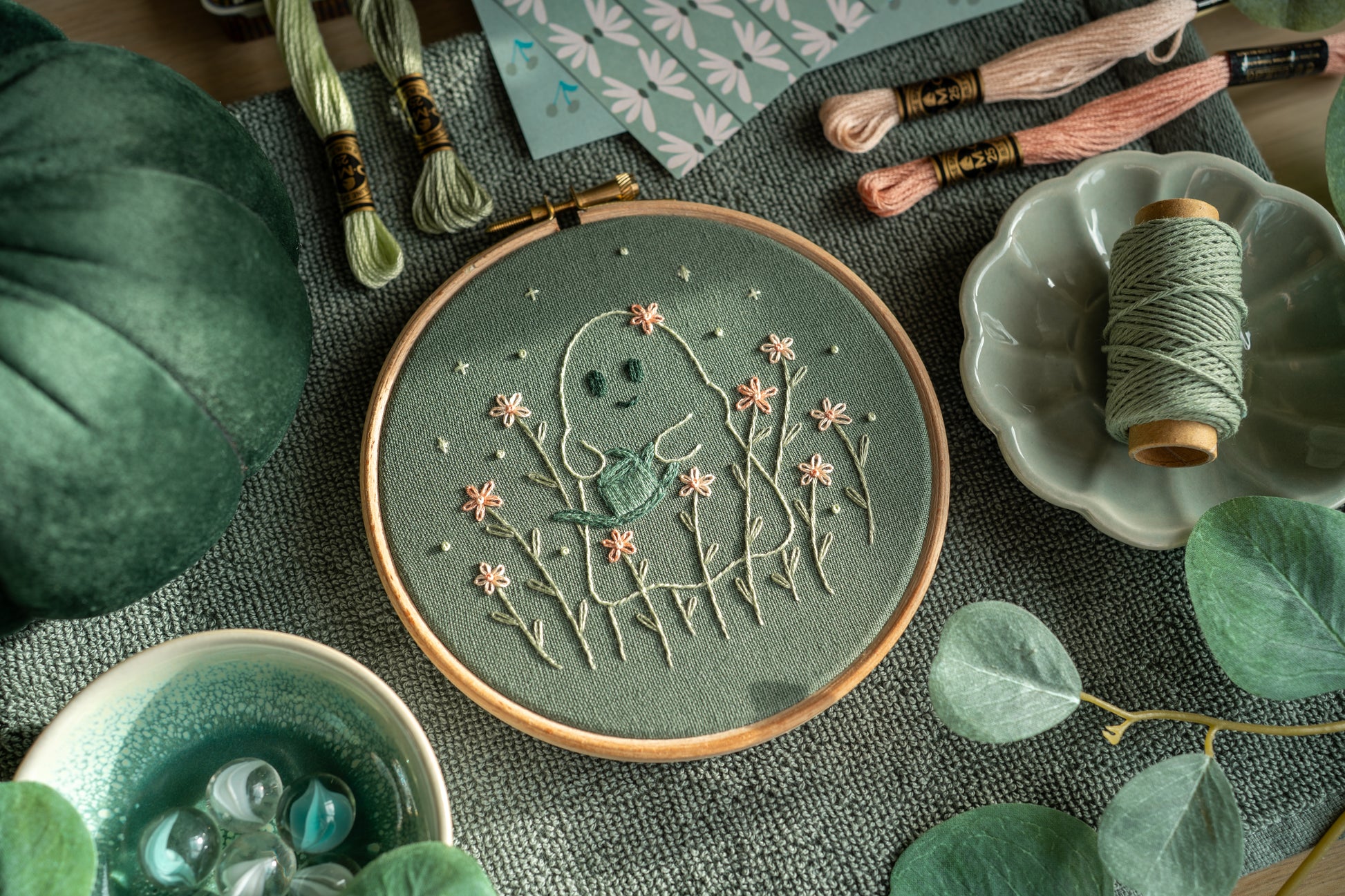 angled view of a hand embroidered ghost gardener design on sage green fabric in a wooden embroidery hoop, surrounded by various green props.