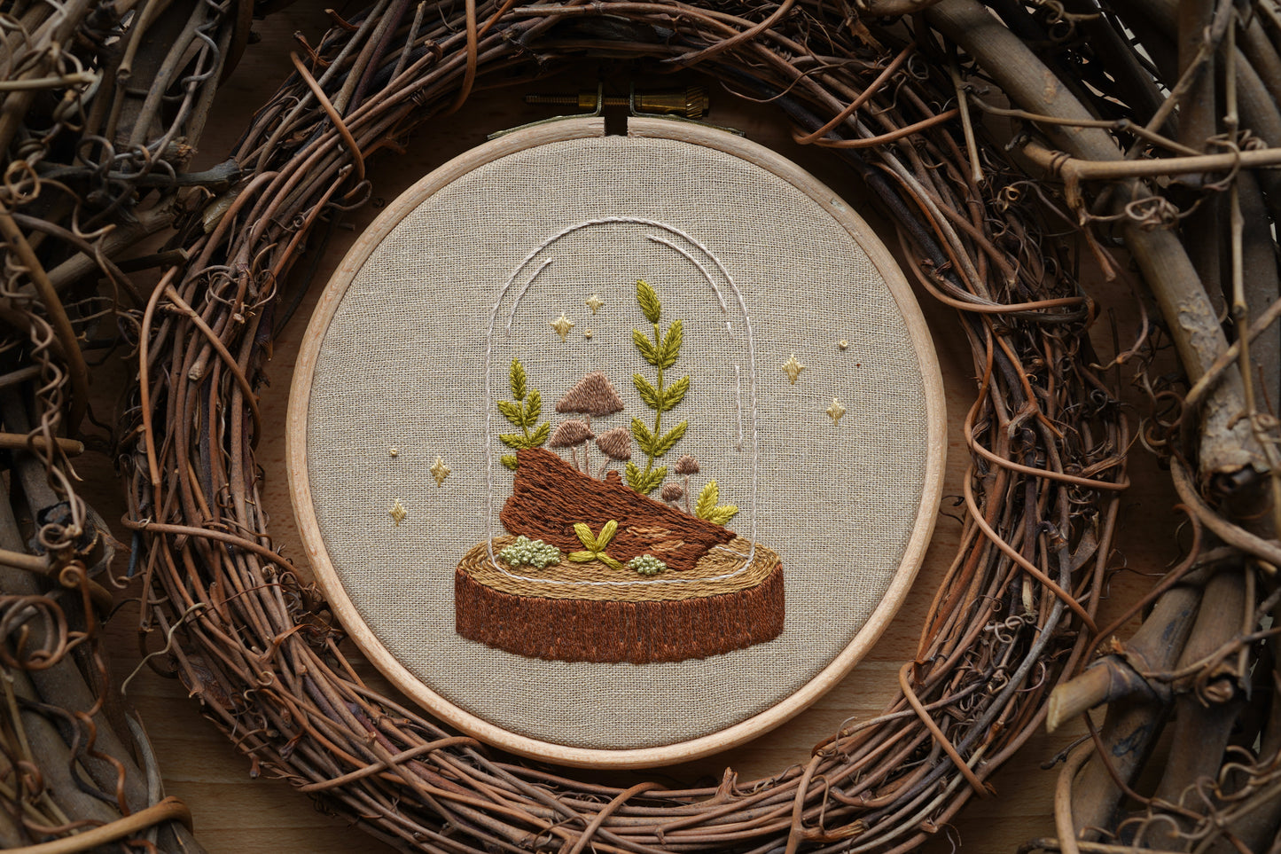 Hand embroidered woodland terrarium design featuring mushrooms and plants on a log in a glass cloche with whimsical sparkles. The hoop is surrounded by twig wreaths.