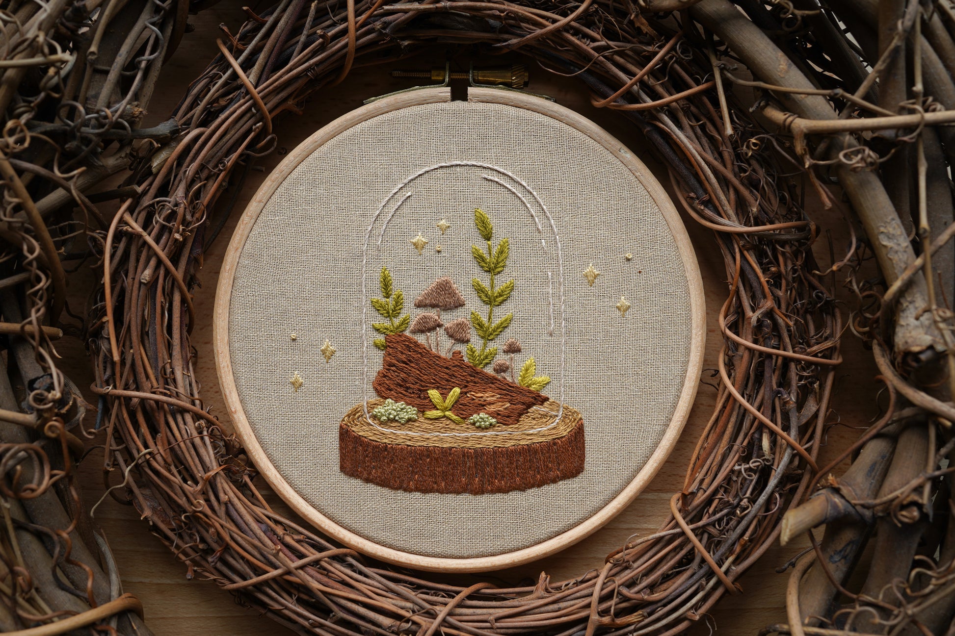 Hand embroidered woodland terrarium design featuring mushrooms and plants on a log in a glass cloche with whimsical sparkles. The hoop is surrounded by twig wreaths.