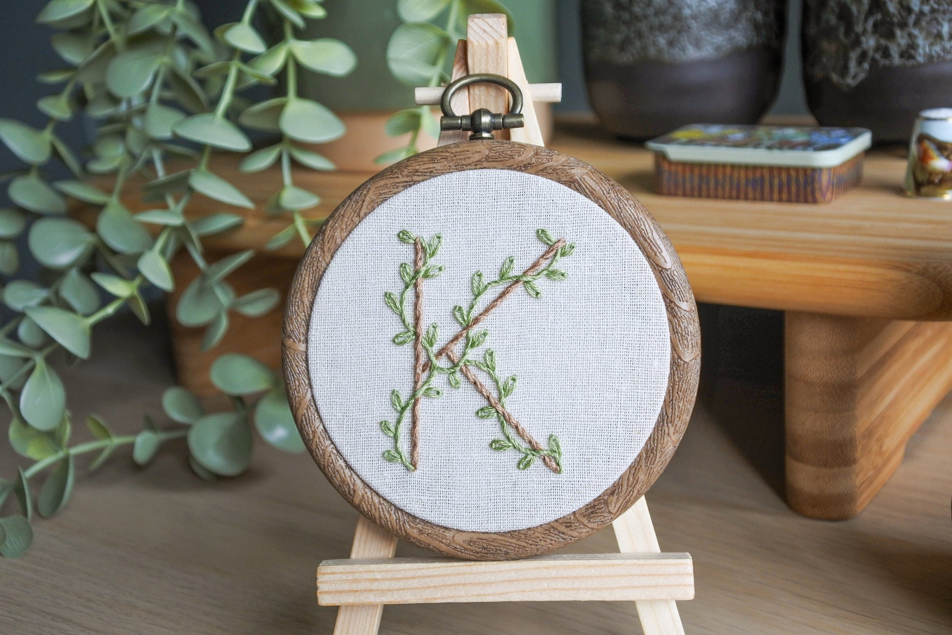 Hand embroidery of botanical letter K with eucalyptus green vines and pale brown stitched letter in a 3-inch wood-effect hoop