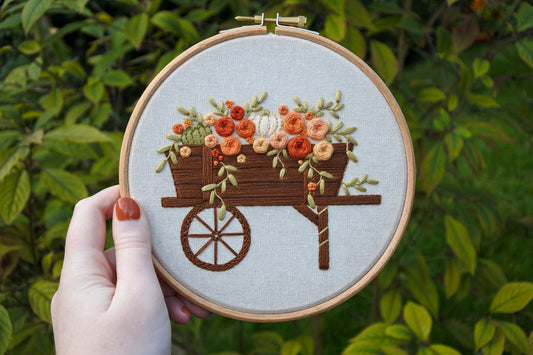 Hand holding hand-embroidered autumn wagon hoop with blurred green leaves in the background