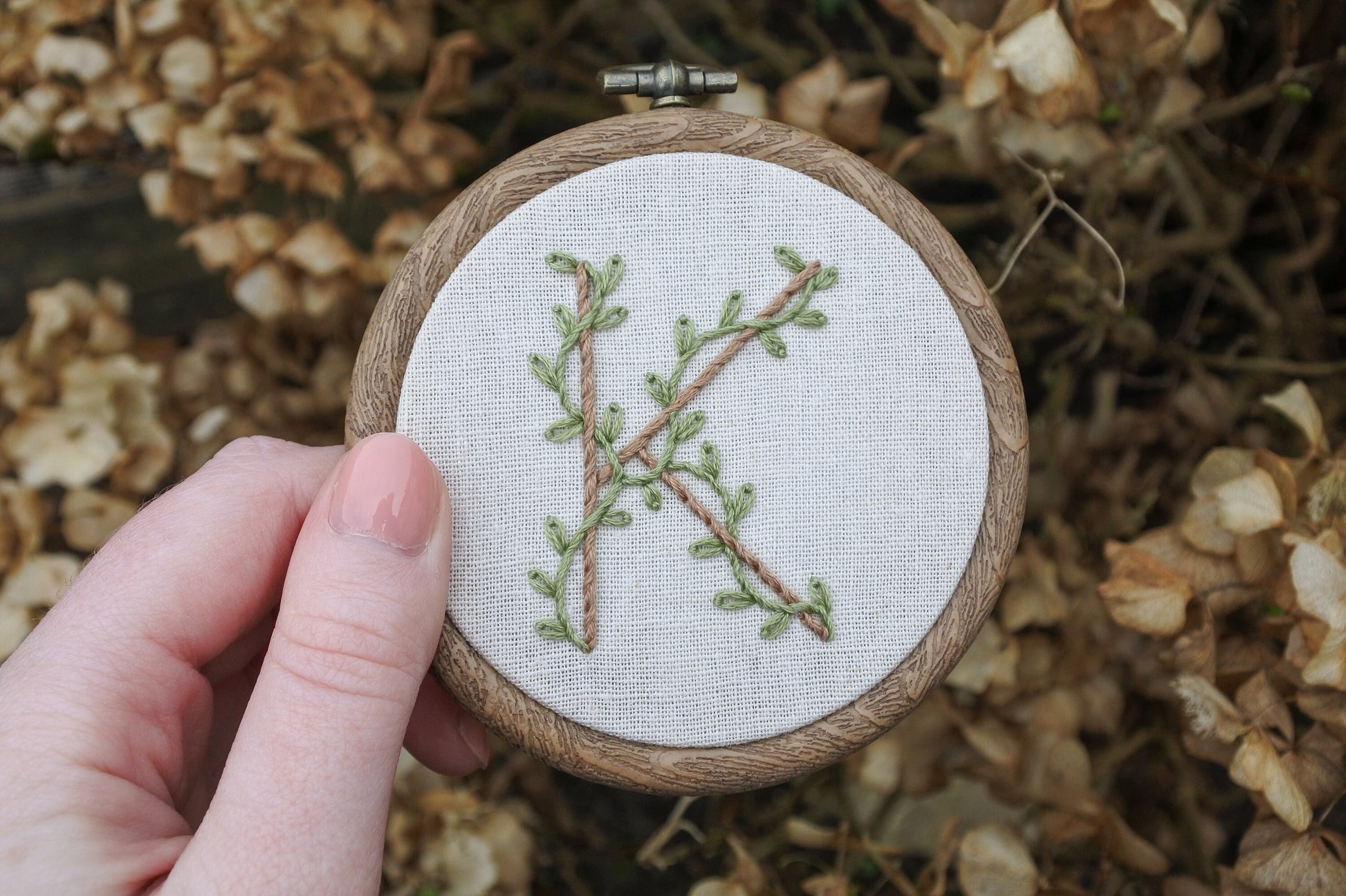 Hand holding botanical letter K embroidery hoop with eucalyptus green vines and pale brown stitched letter on neutral fabric