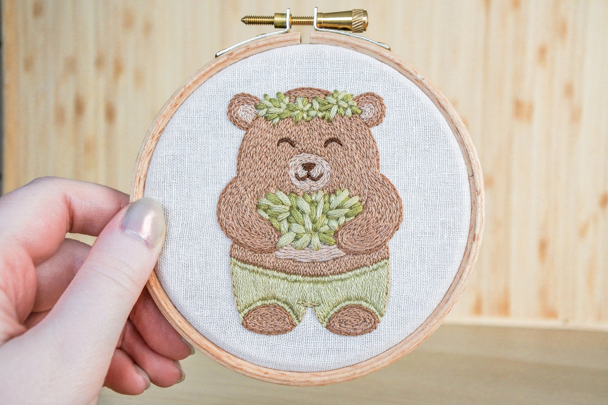 Hand holding a wooden hoop featuring a light brown embroidered bear wearing green shorts and a leaf crown, holding a basket of leaves