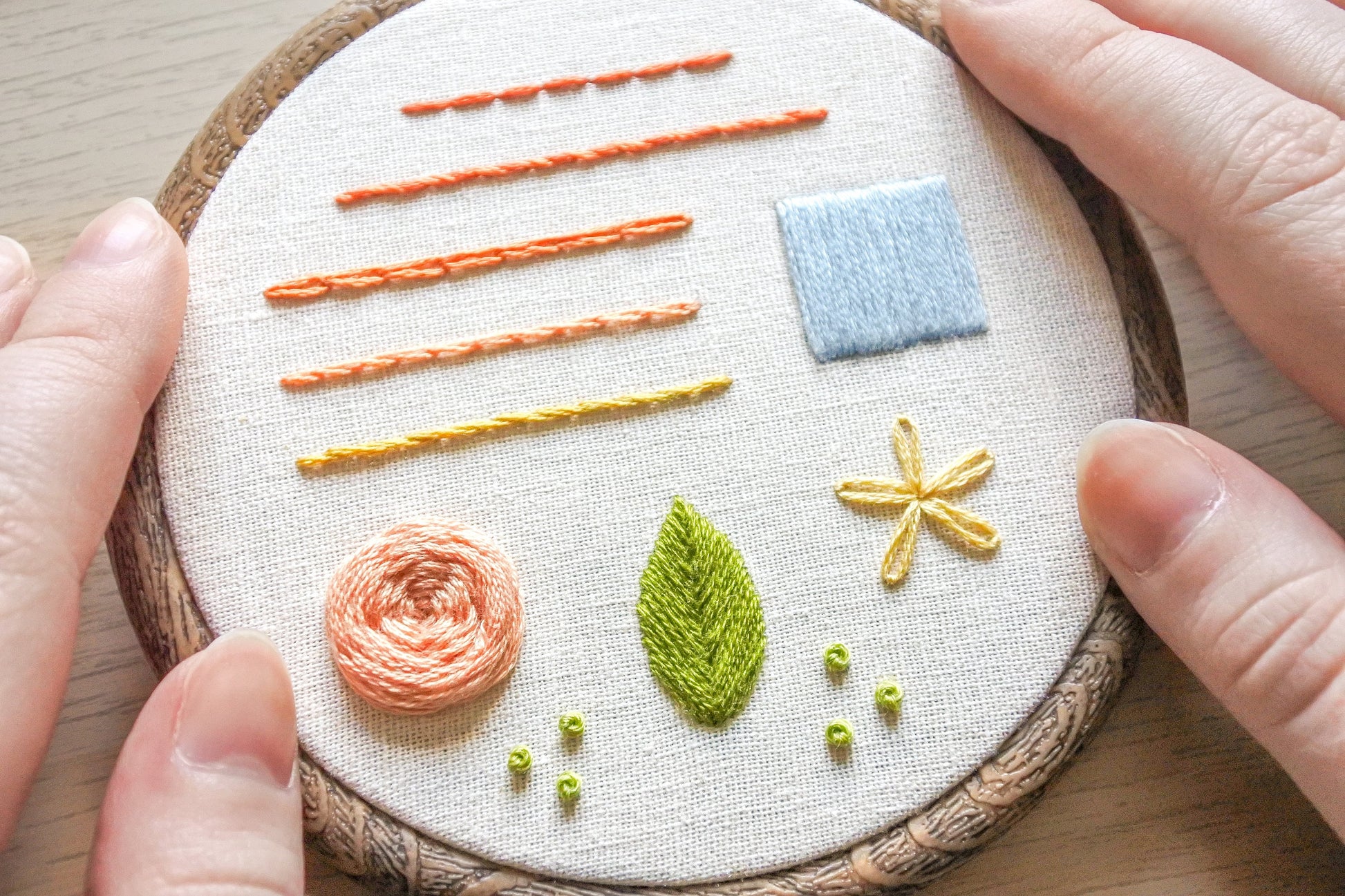 Hands holding a finished embroidery hoop featuring a stitch sampler with 10 essential beginner stitches on neutral fabric