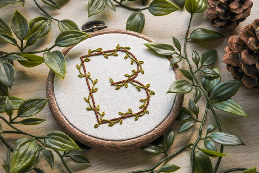 Hand-embroidered botanical letter 'B' hoop, with jungle green leafy vines and dark brown thread on light neutral fabric. The wood effect hoop is surrounded by vine leaves.