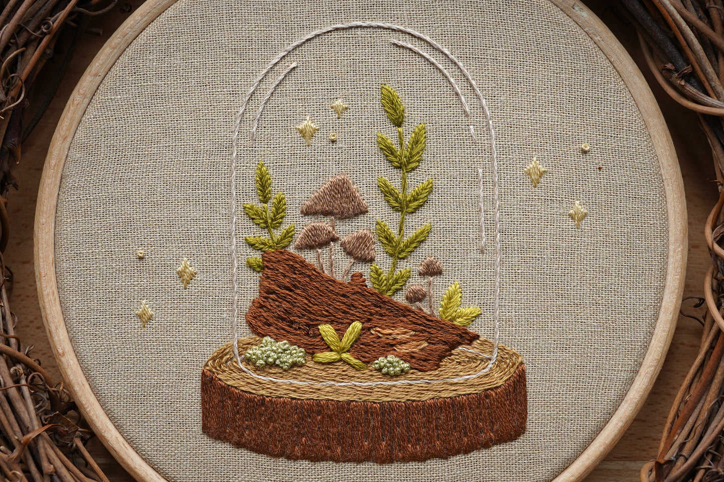Closeup of embroidery featuring a  woodland terrarium with plants and mushrooms growing on a log in a wooden hoop.
