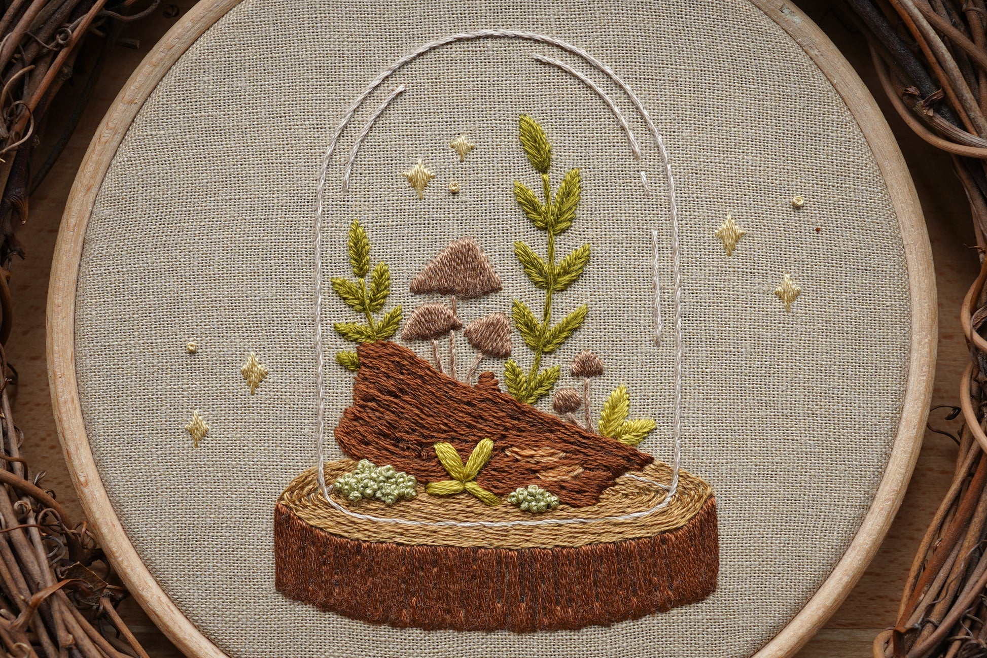 Closeup of embroidery featuring a  woodland terrarium with plants and mushrooms growing on a log in a wooden hoop.