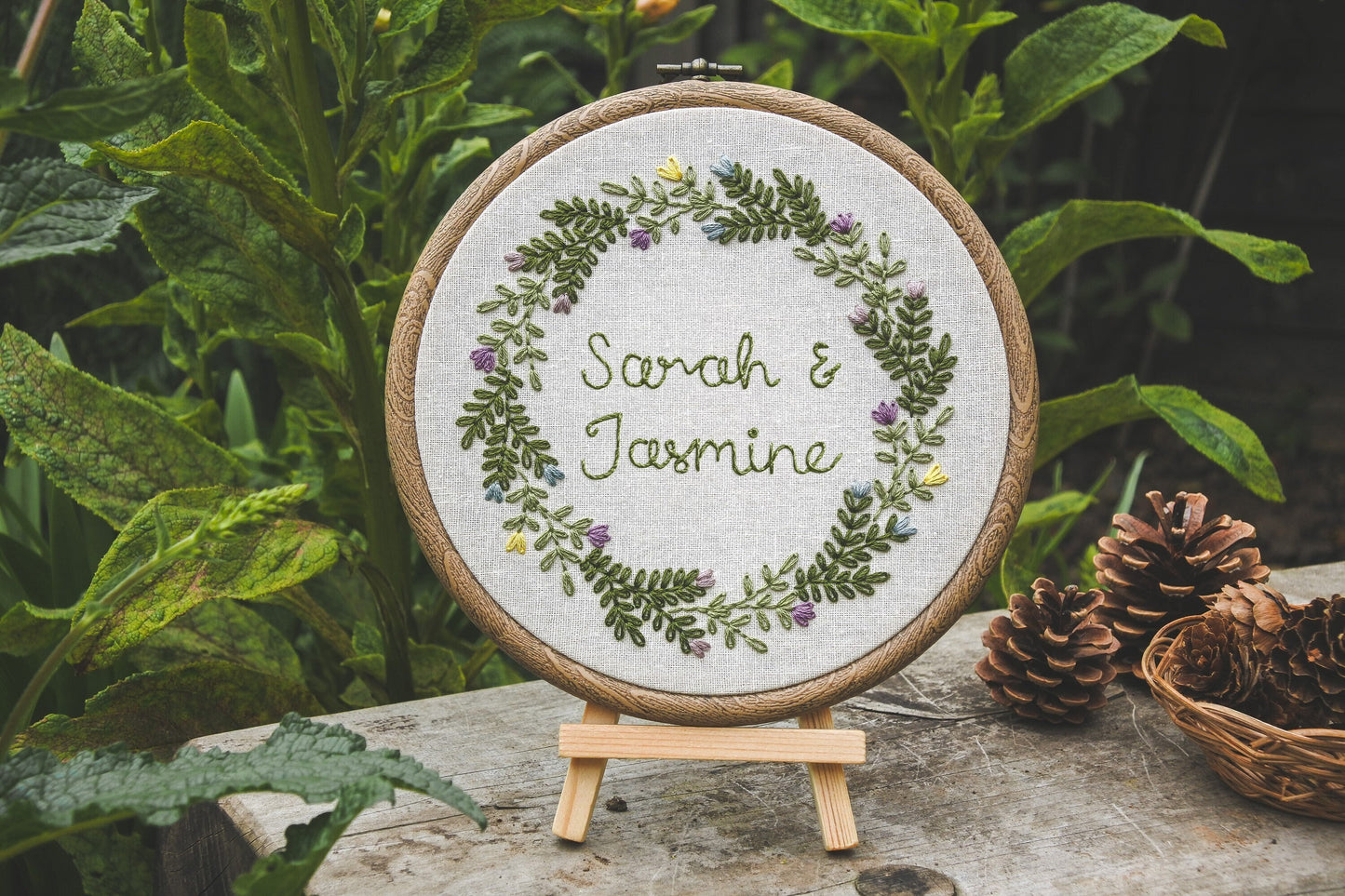 Personalised spring wedding embroidery hoop with the names Sarah and Jasmine, displayed on a wooden easel surrounded by greenery.