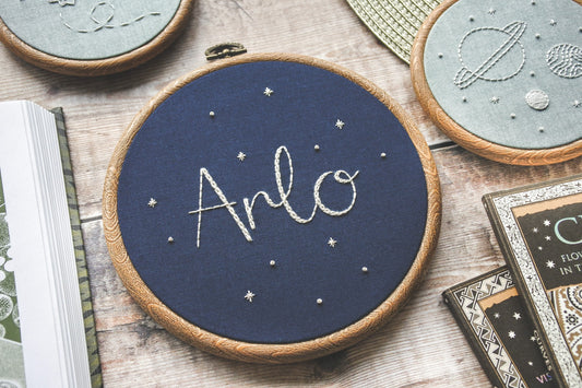 Hand embroidered name hoop with the name Arlo stitched in white thread on navy fabric, framed in a 6-inch wood-effect hoop