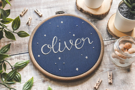 Hand embroidered name 'Oliver' surrounded by white stars on navy blue fabric in a wood-effect hoop