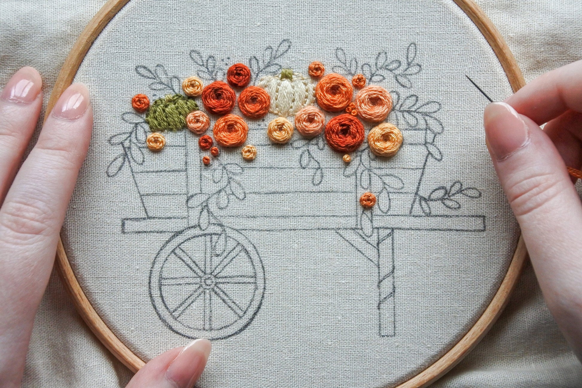 Hand holding a needle and thread while stitching flowers and pumpkins on an autumn wagon embroidery pattern