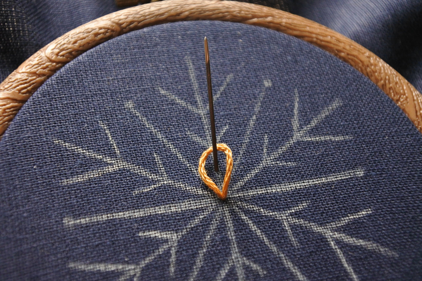 Progress photo of a needle stitching a gold chain stitch onto navy blue fabric