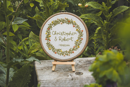 Personalised summer wedding embroidery hoop with “Christopher and Robert” and their wedding date, displayed outdoors on a wooden easel surrounded by greenery