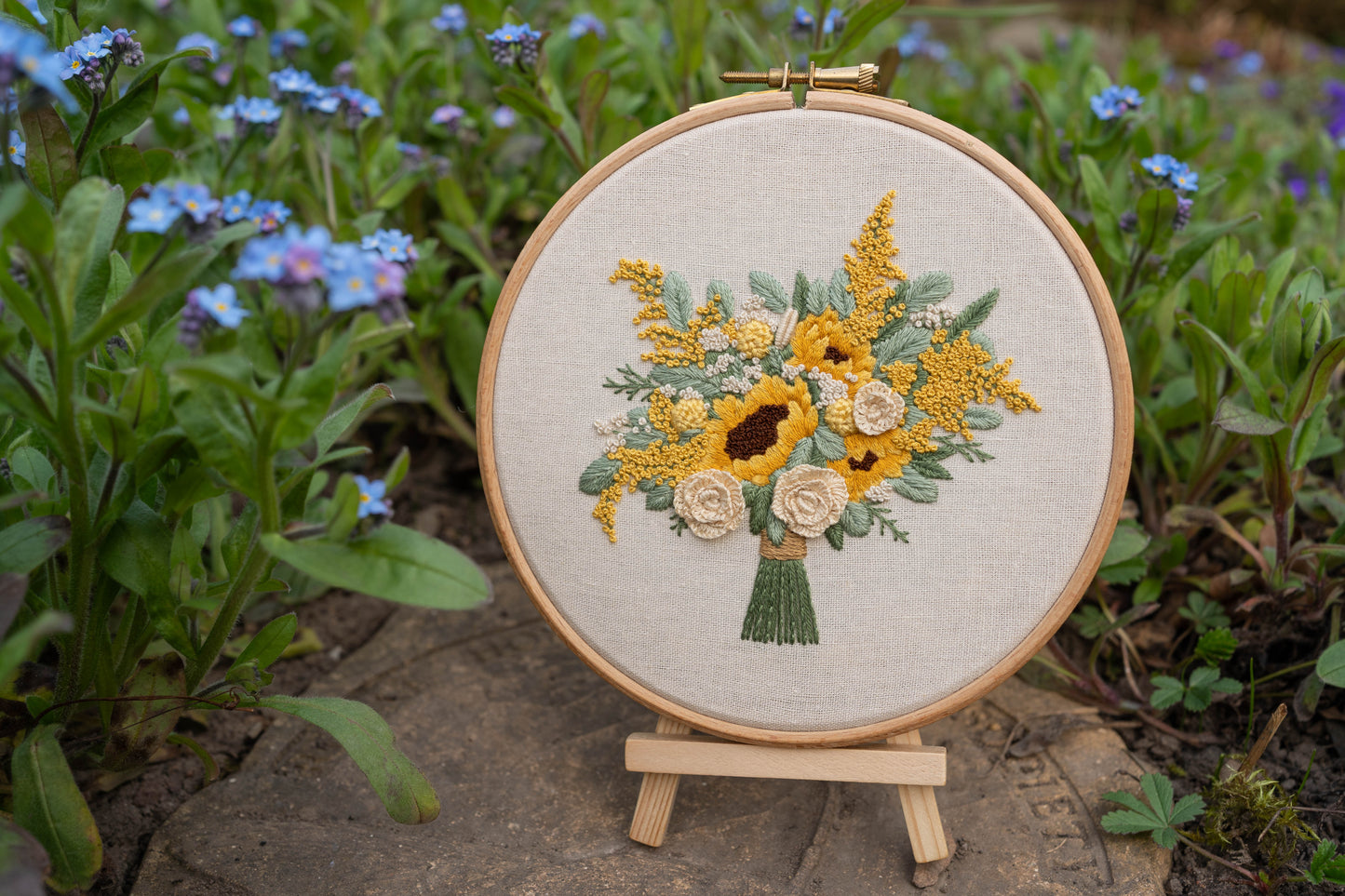 Custom hand-embroidered sunflower bouquet hoop displayed on a small wooden easel surrounded by plants