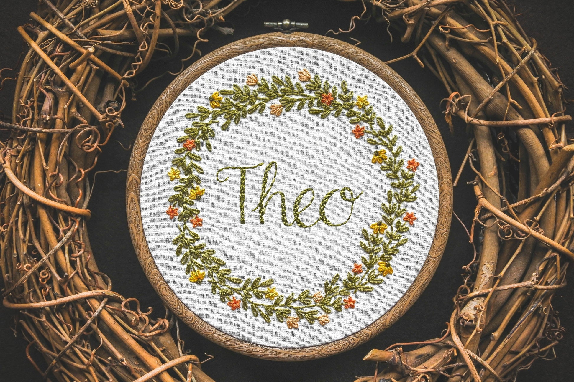 Hand embroidered summer wreath name hoop with the name Theo stitched in the centre, featuring bright floral details and warm green leaves.