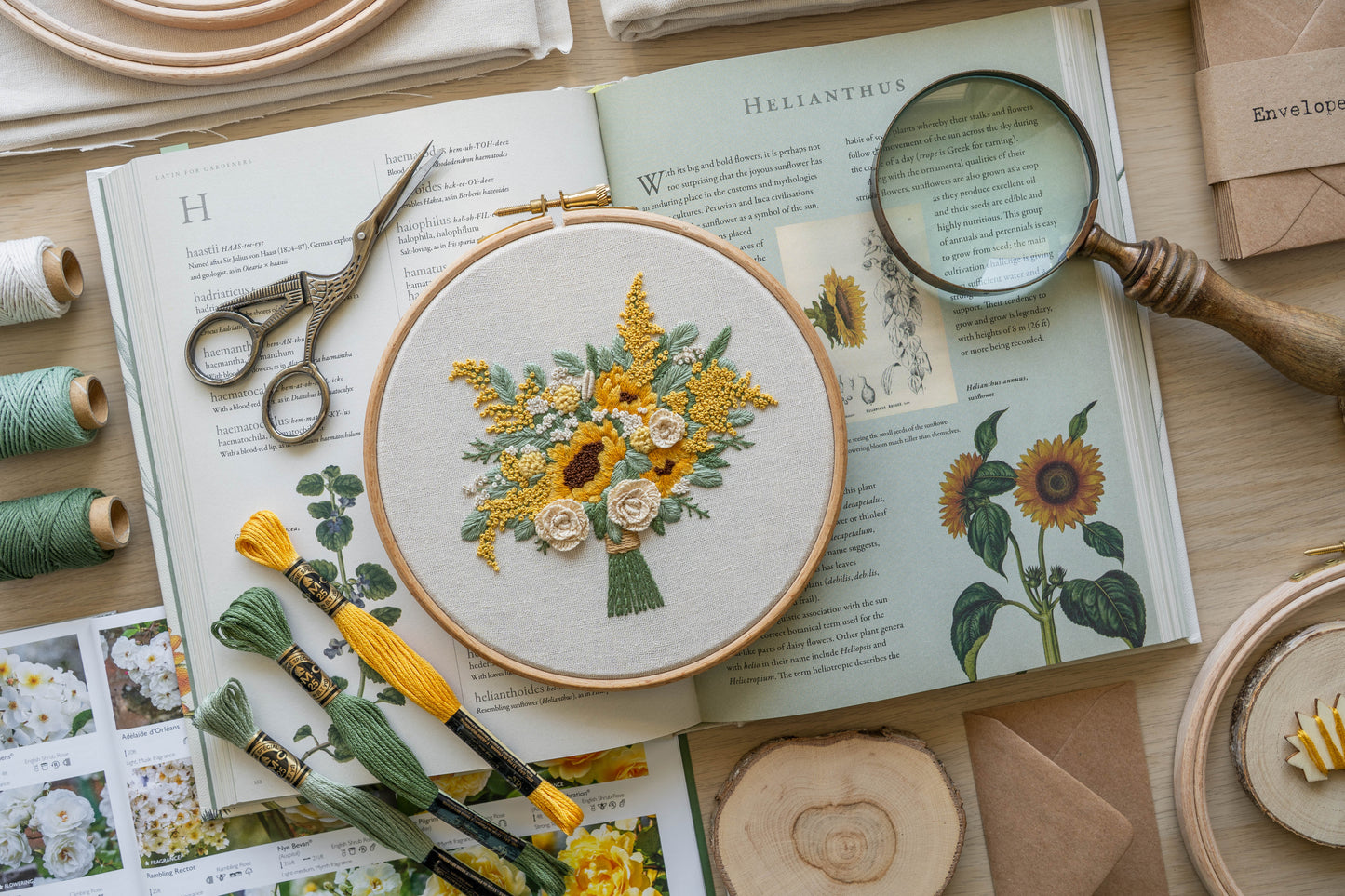 Decorative flatlay of a hand embroidered floral bouquet featuring sunflowers, cream roses and eucalyptus foliage, in a wooden hoop set on top of a botanical book
