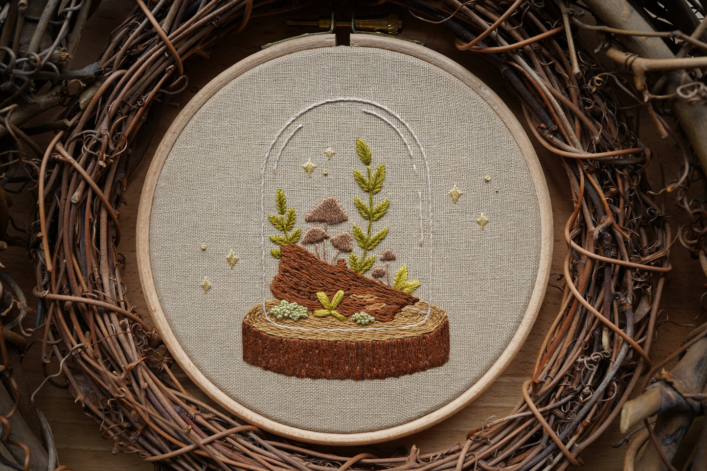 Closeup of embroidery featuring a whimsical woodland terrarium with plants and mushrooms growing on a log in a wooden hoop. Sparkles surround the terrarium.