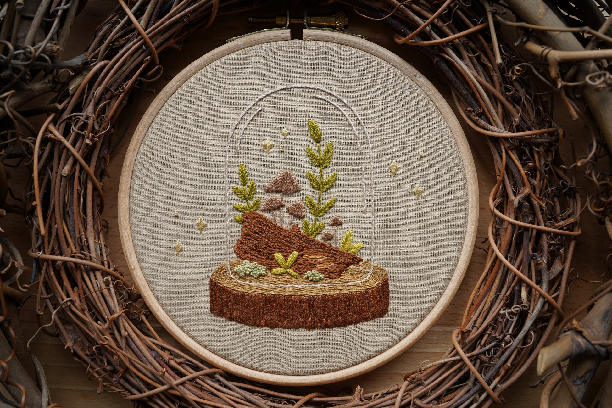 Closeup of embroidery featuring a whimsical woodland terrarium with plants and mushrooms growing on a log in a wooden hoop. Sparkles surround the terrarium.