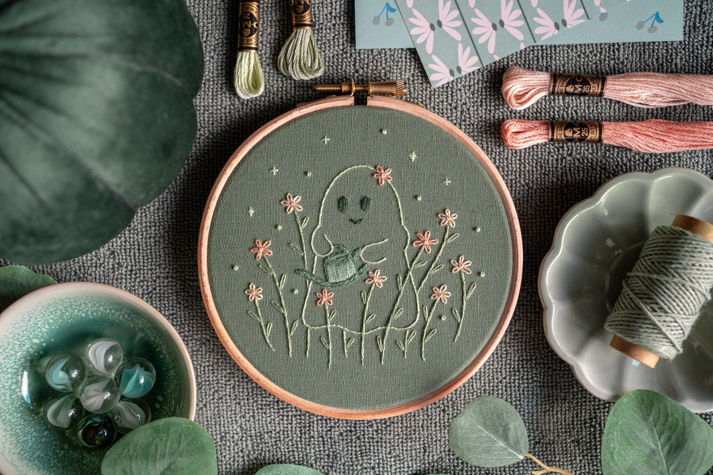 Whimsical ghost gardener hand embroidery art in a wooden embroidery hoop surrounded by various props.