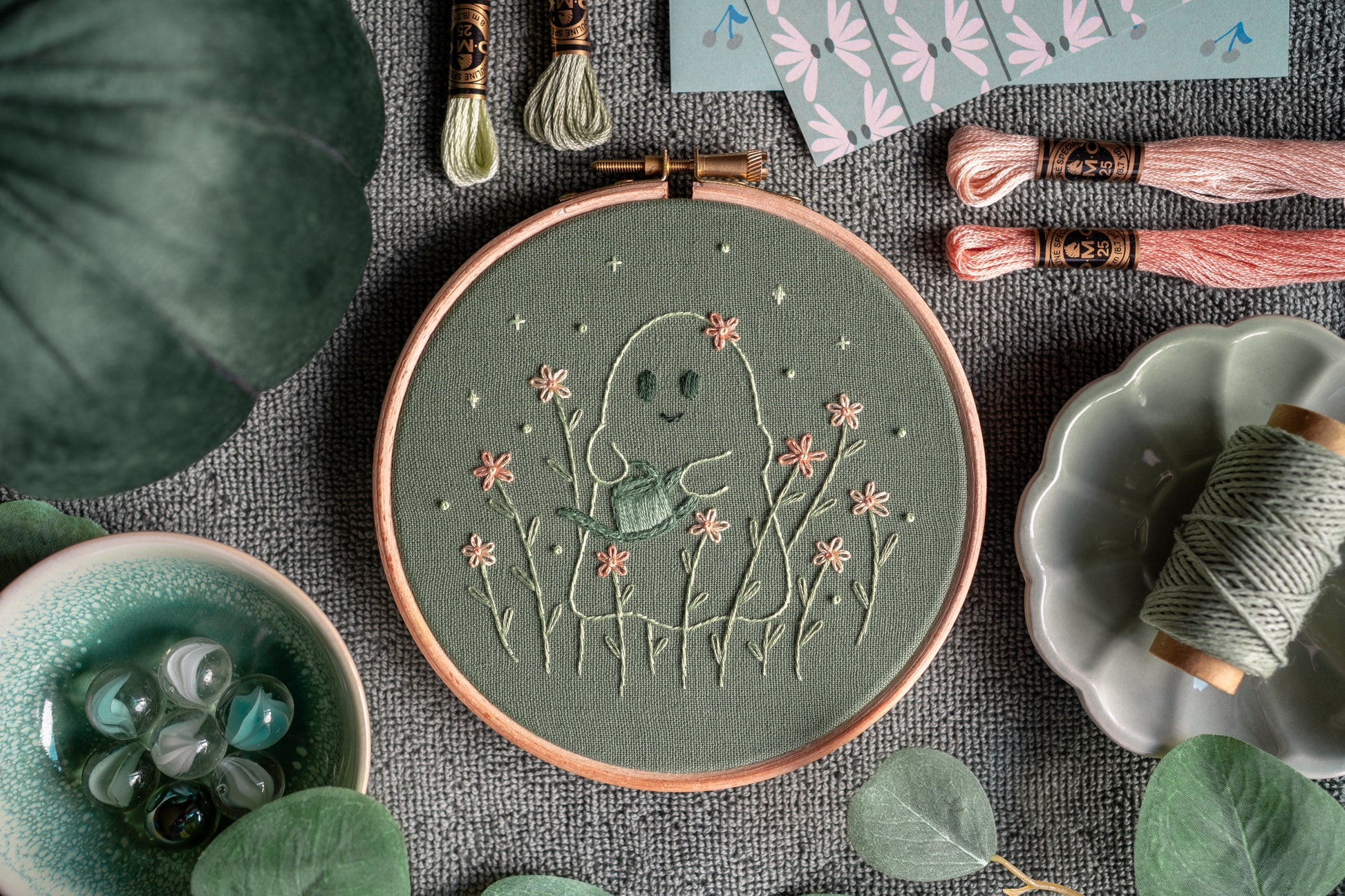 Whimsical ghost gardener hand embroidery art in a wooden embroidery hoop surrounded by various props.