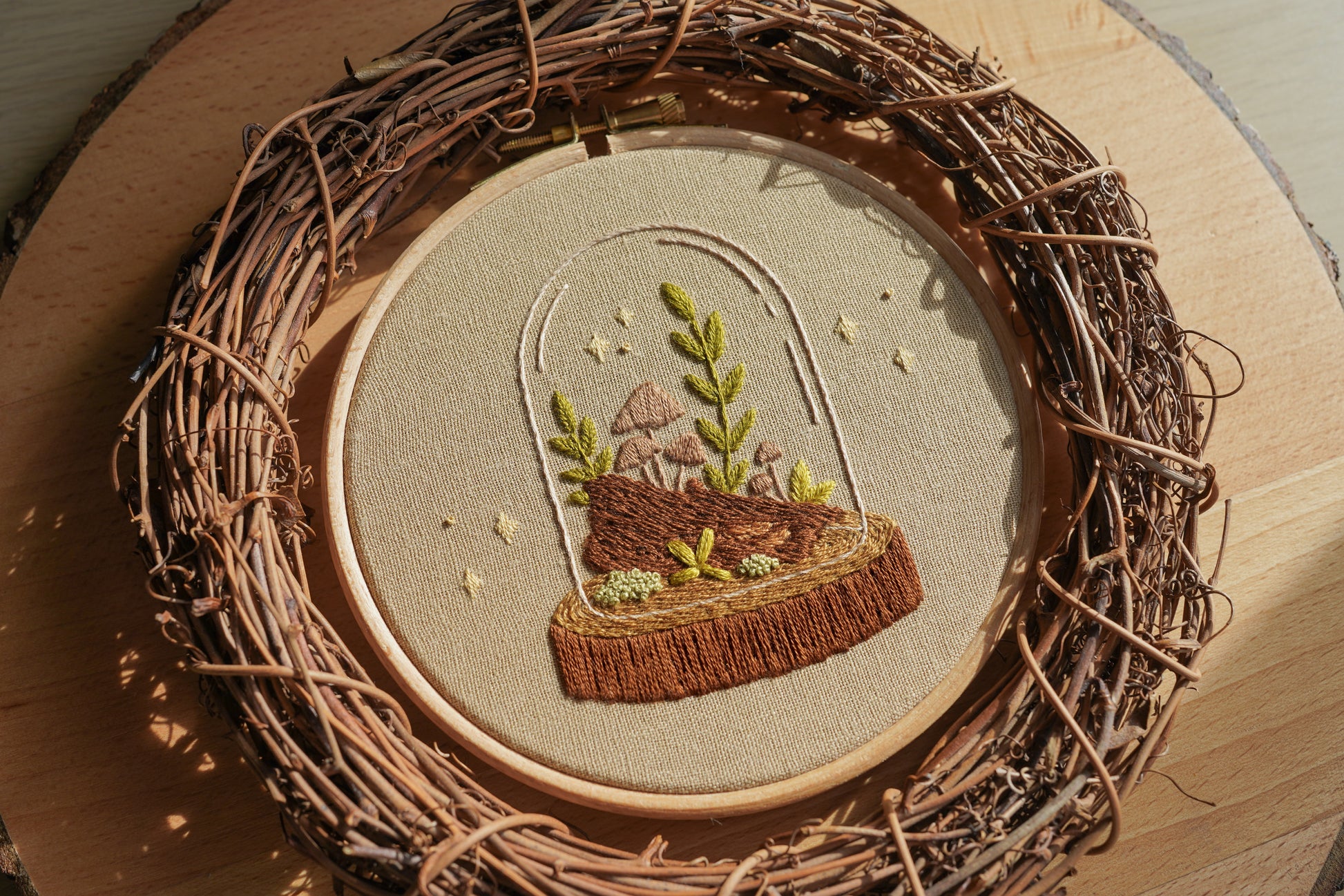 Embroidery of a woodland terrarium scene in a wooden hoop with a natural, rustic background.