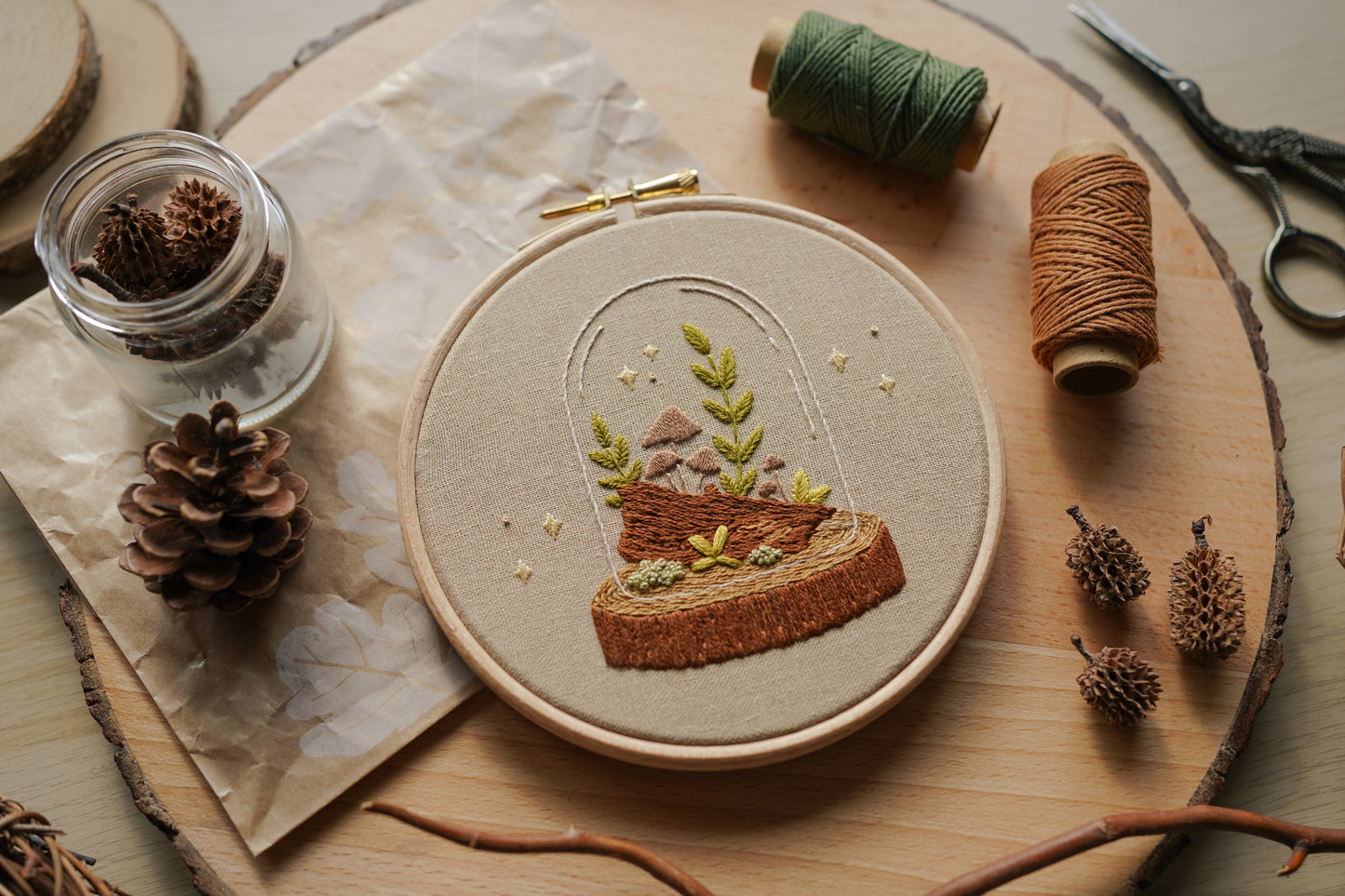 Embroidery hoop with nature-themed woodland terrarium fungi design on a wooden surface with natural elements.