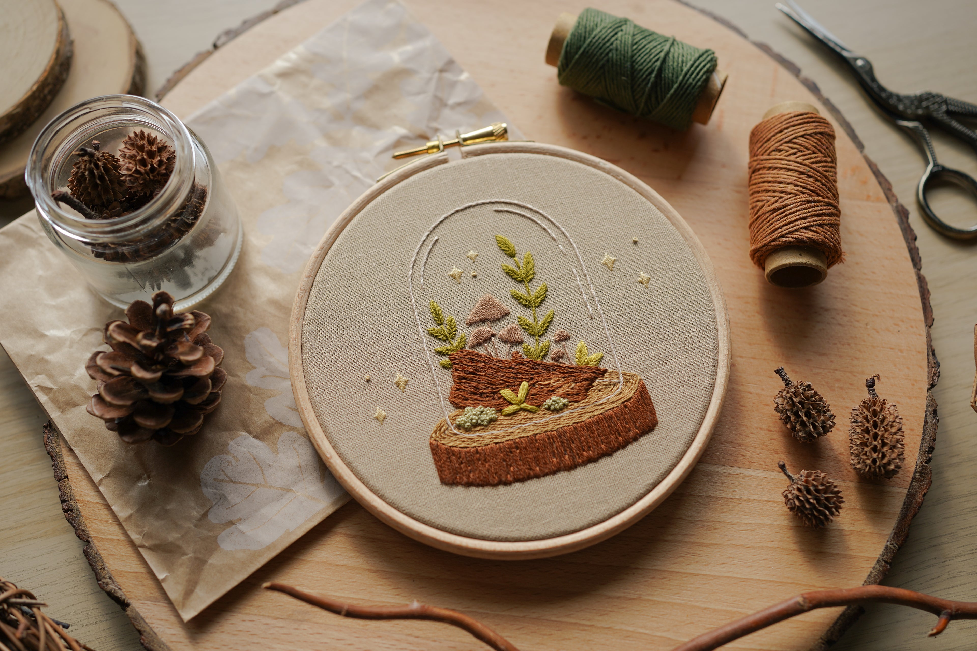 Embroidery hoop with nature-themed woodland terrarium fungi design on a wooden surface with natural elements.
